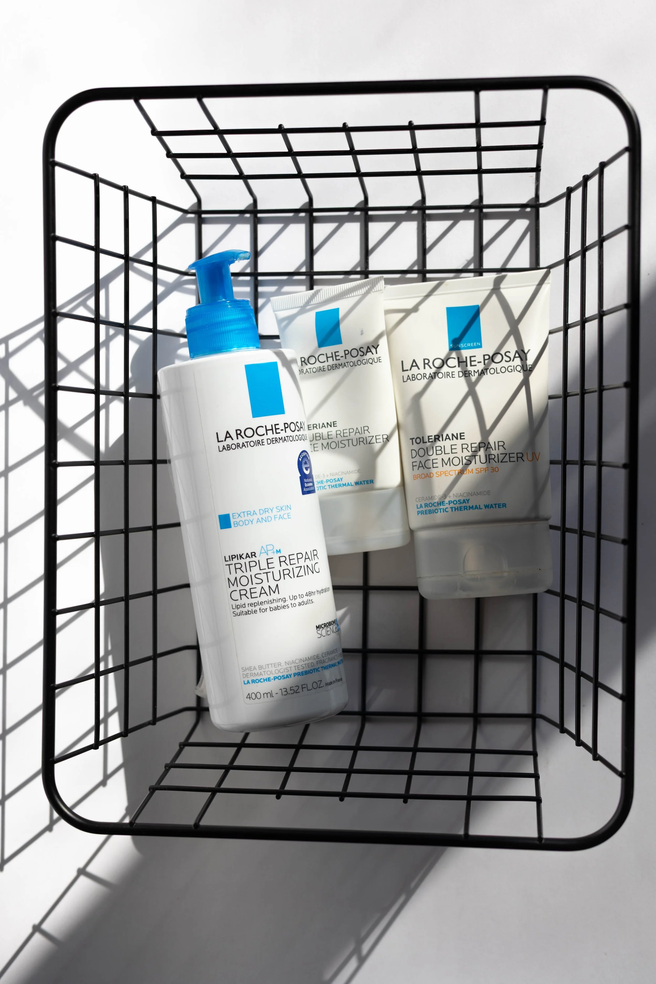 A black metal wire basket holding three tubes and a bottle of La Roche-Posay skincare products, casts shadows on a white surface.
