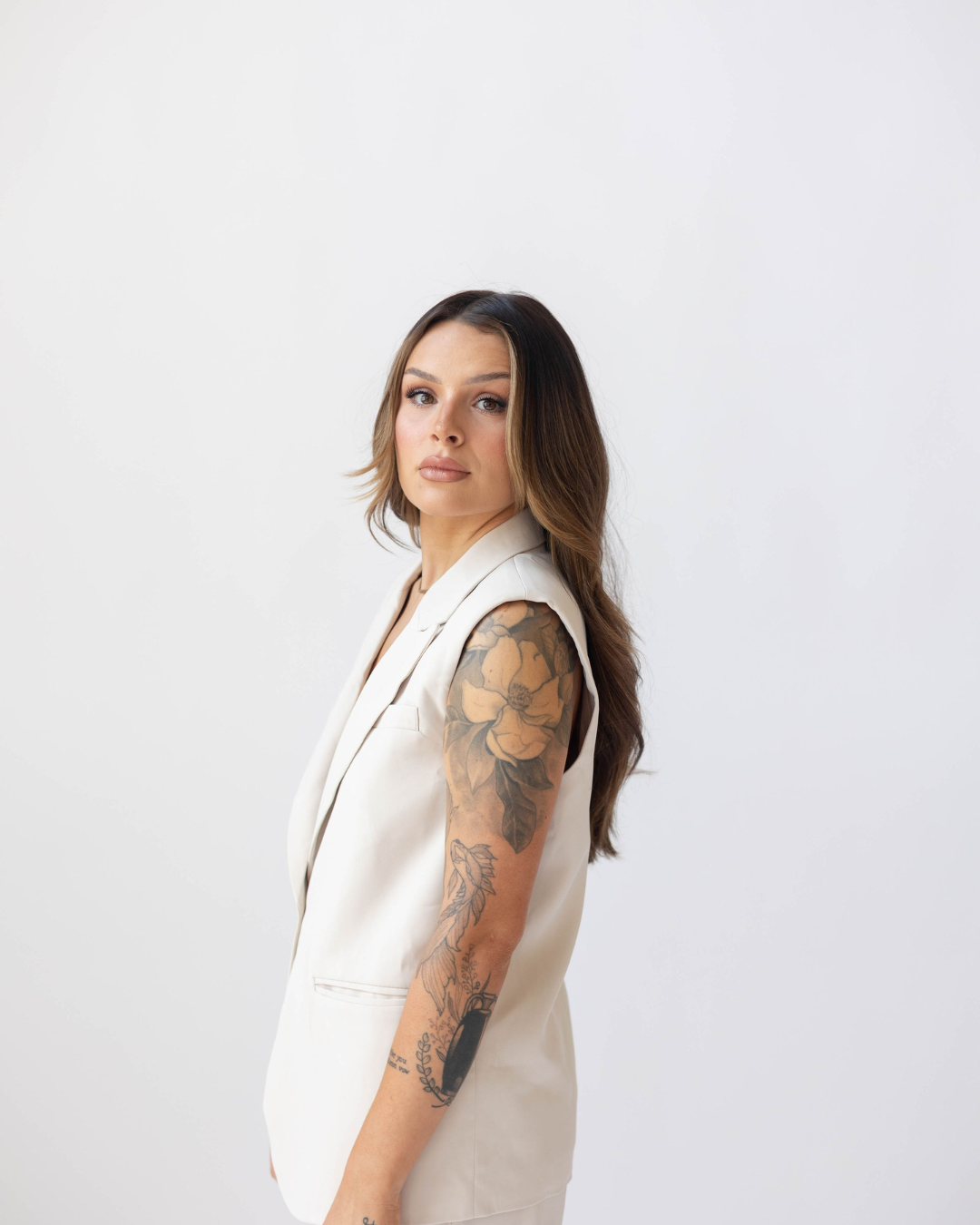 A woman with long brown hair, wearing a sleeveless white blazer, stands against a plain white background, looking at the camera with a neutral expression.