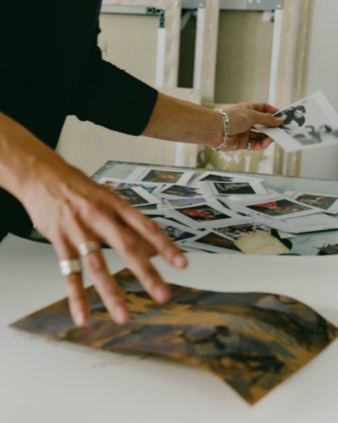 Person in black sleeve handling printed photographs on a white table, with more photos scattered across the surface, in a room with unfinished walls.