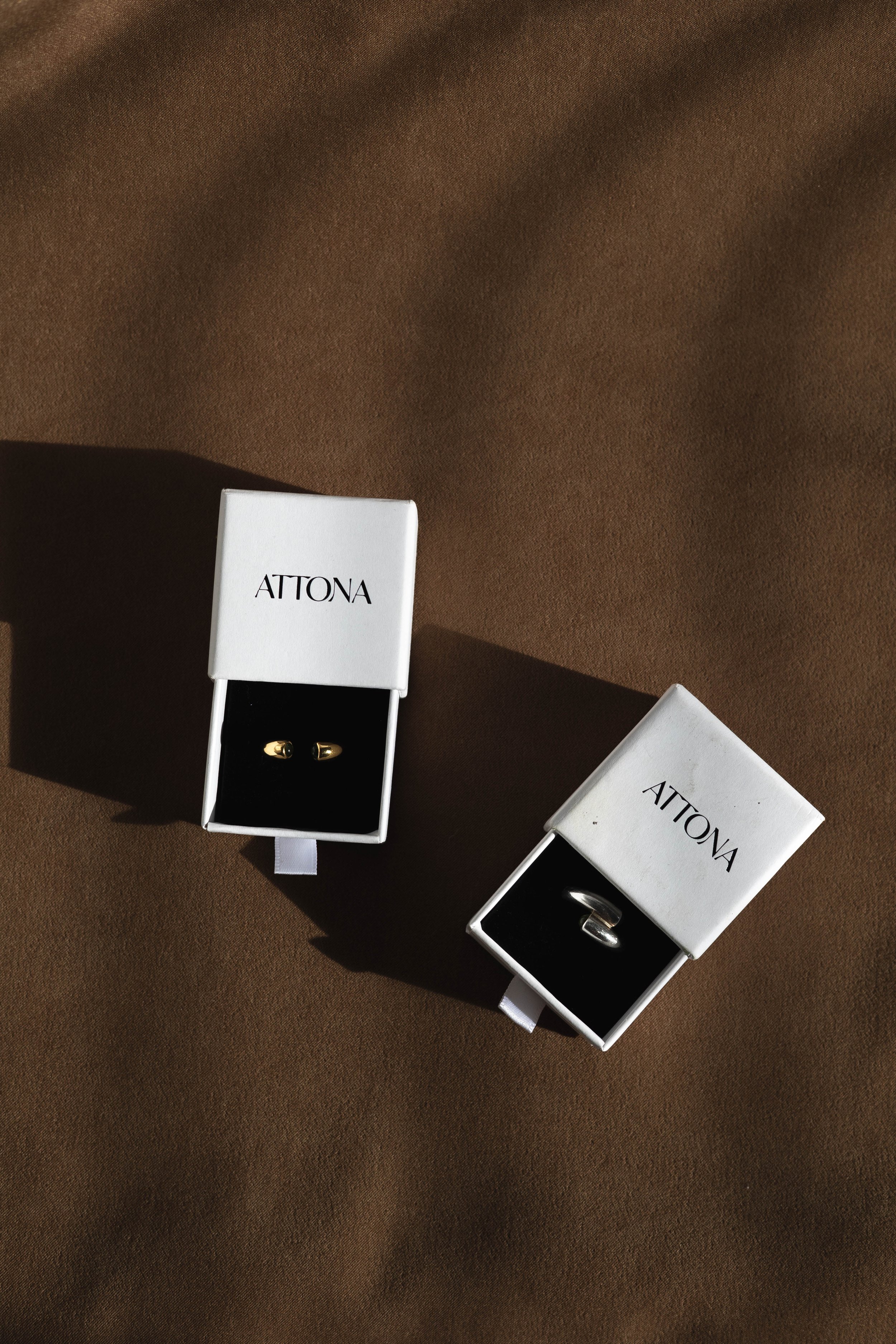 Two jewelry boxes labeled ATTONA, each containing a pair of gold earrings, placed on a brown surface with shadows.