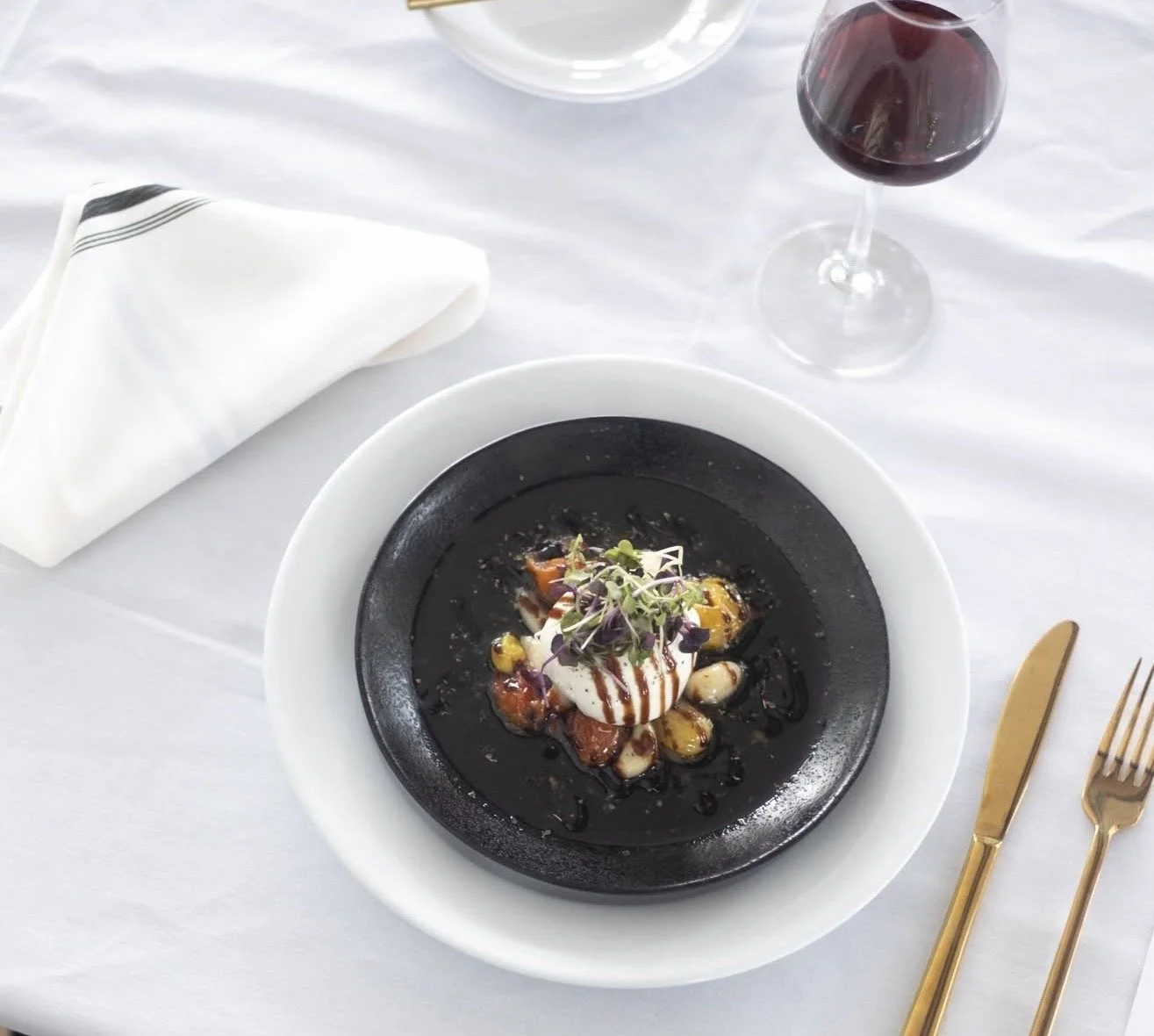 A plated dish with microgreens, sauce, and dollops of cream on a black bowl on white tablecloth, with a glass of red wine, a white napkin, and gold-colored flatware.