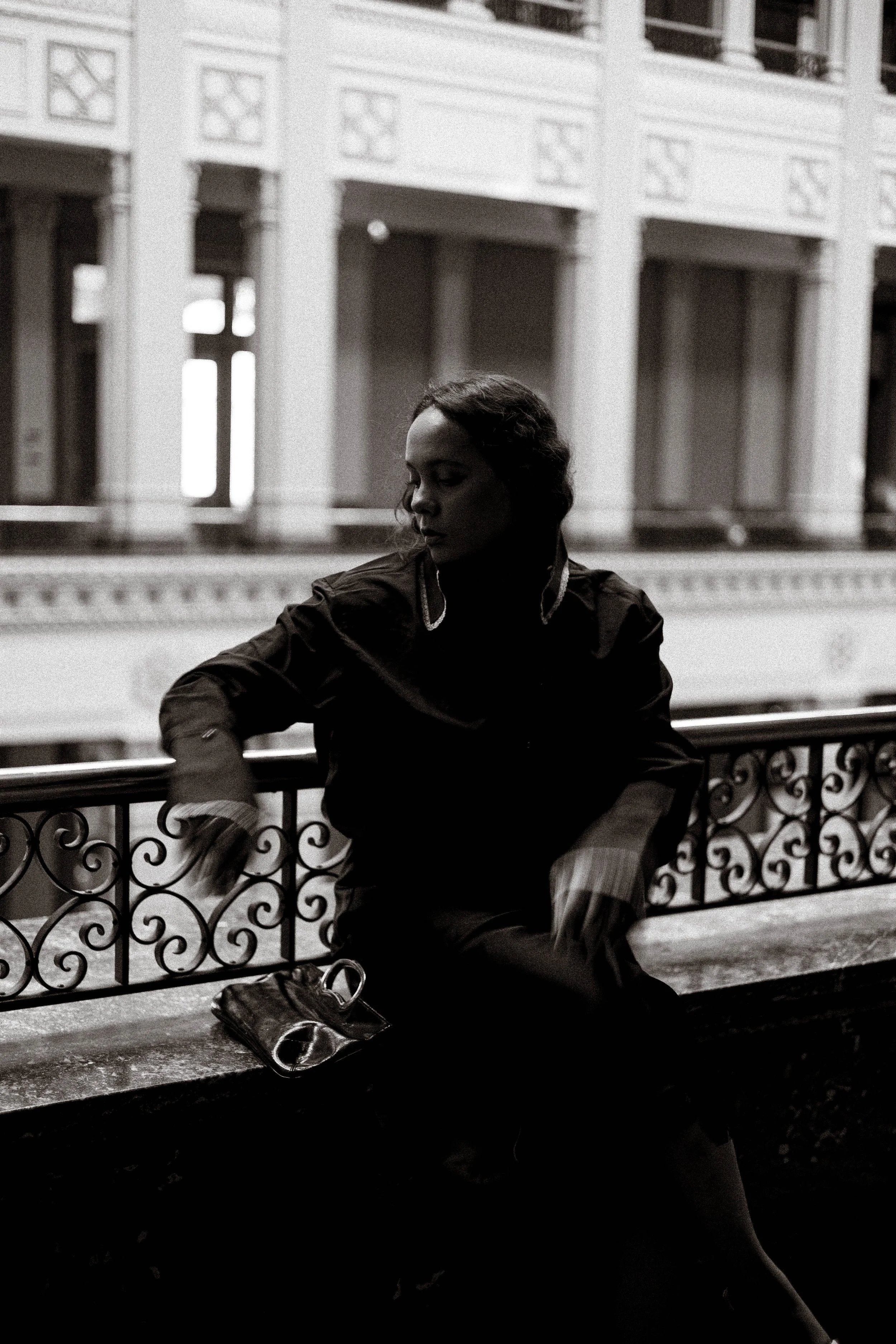 A woman sitting on a bench outdoors, looking down with closed eyes, in front of a building with ornate architecture in black and white.
