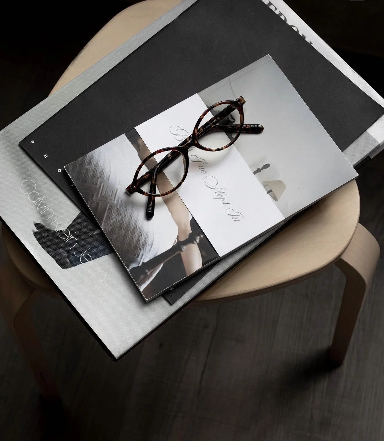 A pair of tortoise shell glasses resting on a stack of books and magazines on a wooden chair.