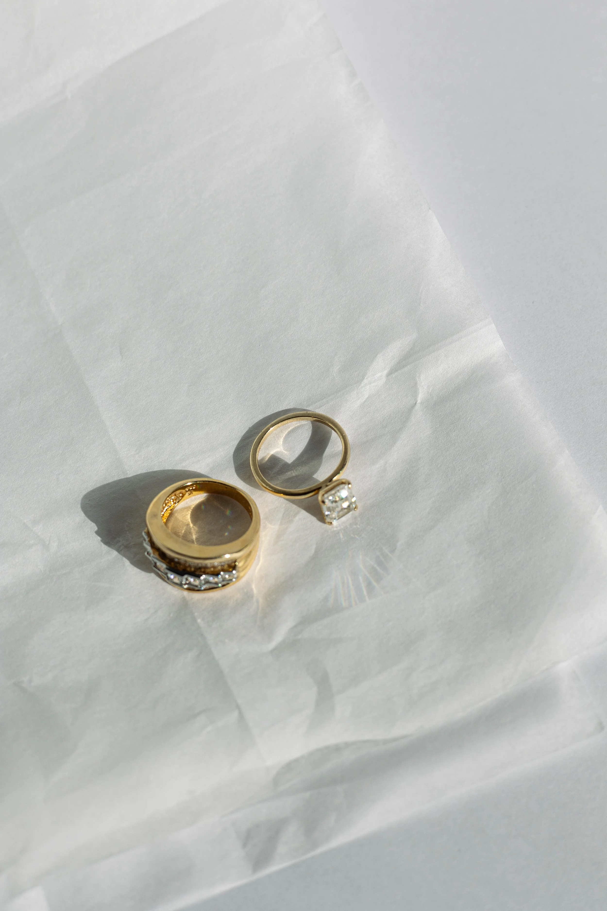 Two gold rings with gemstones resting on white tissue paper, creating shadows and reflections.