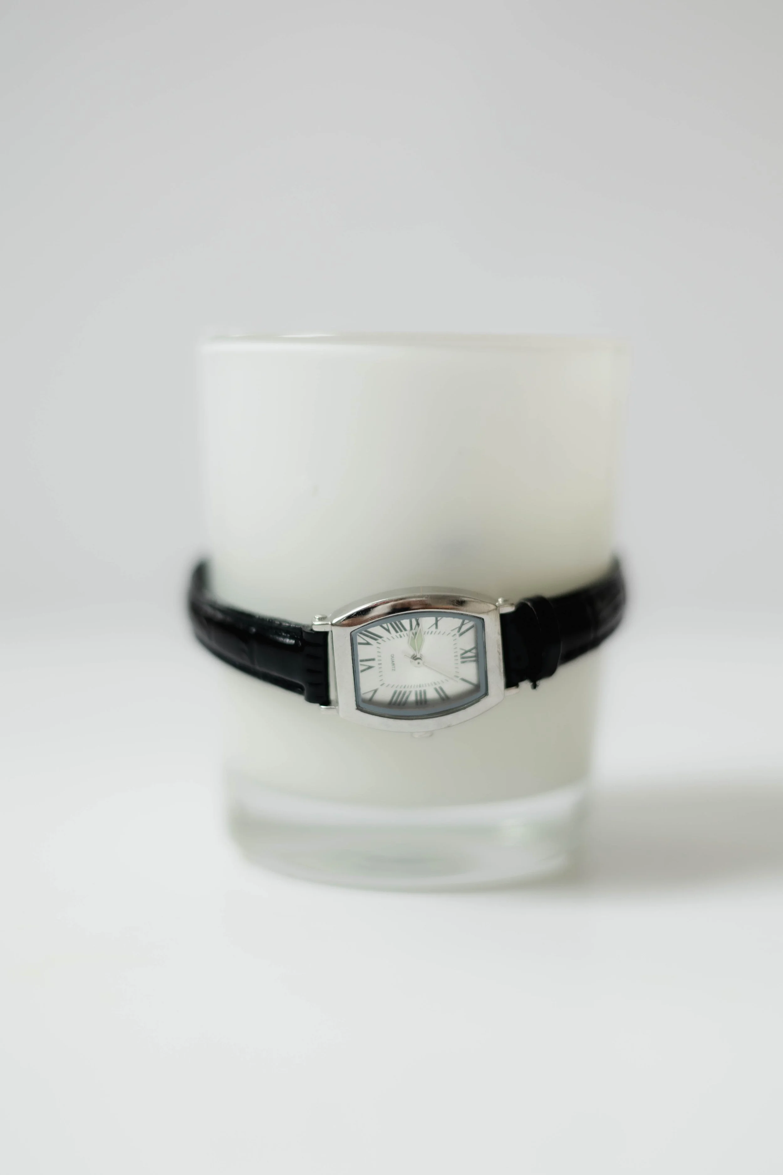 A rectangular wristwatch with a white face, Roman numeral markers, and a black leather strap, placed over a white cylindrical container against a plain white background.