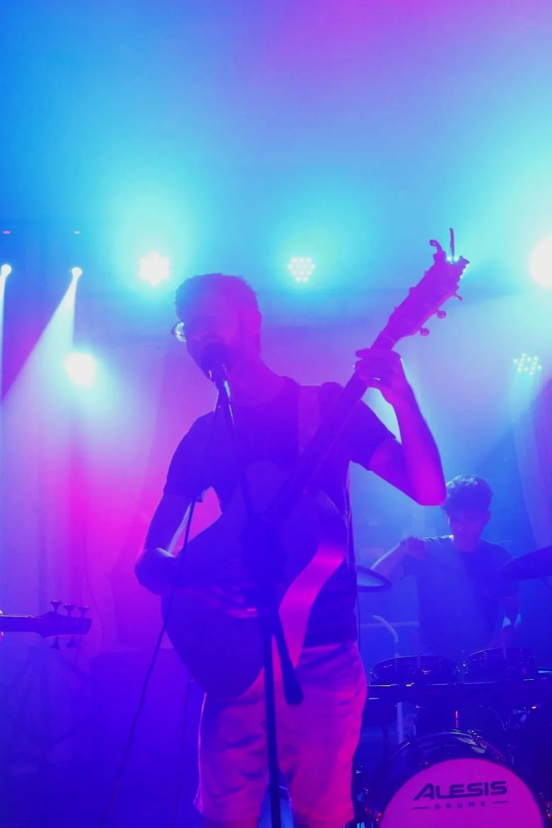 A person playing guitar and singing into a microphone on stage, with colorful purple and blue stage lights, and a drummer visible in the background.