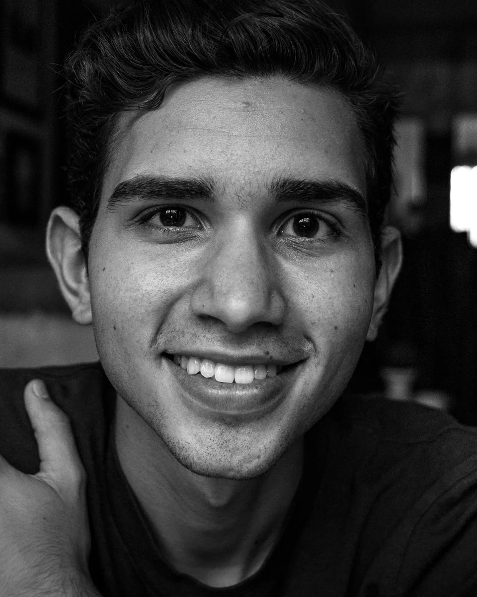 Black and white close-up photo of a smiling young man with short hair, visible teeth, and one hand touching his shoulder, indoors with blurred background.