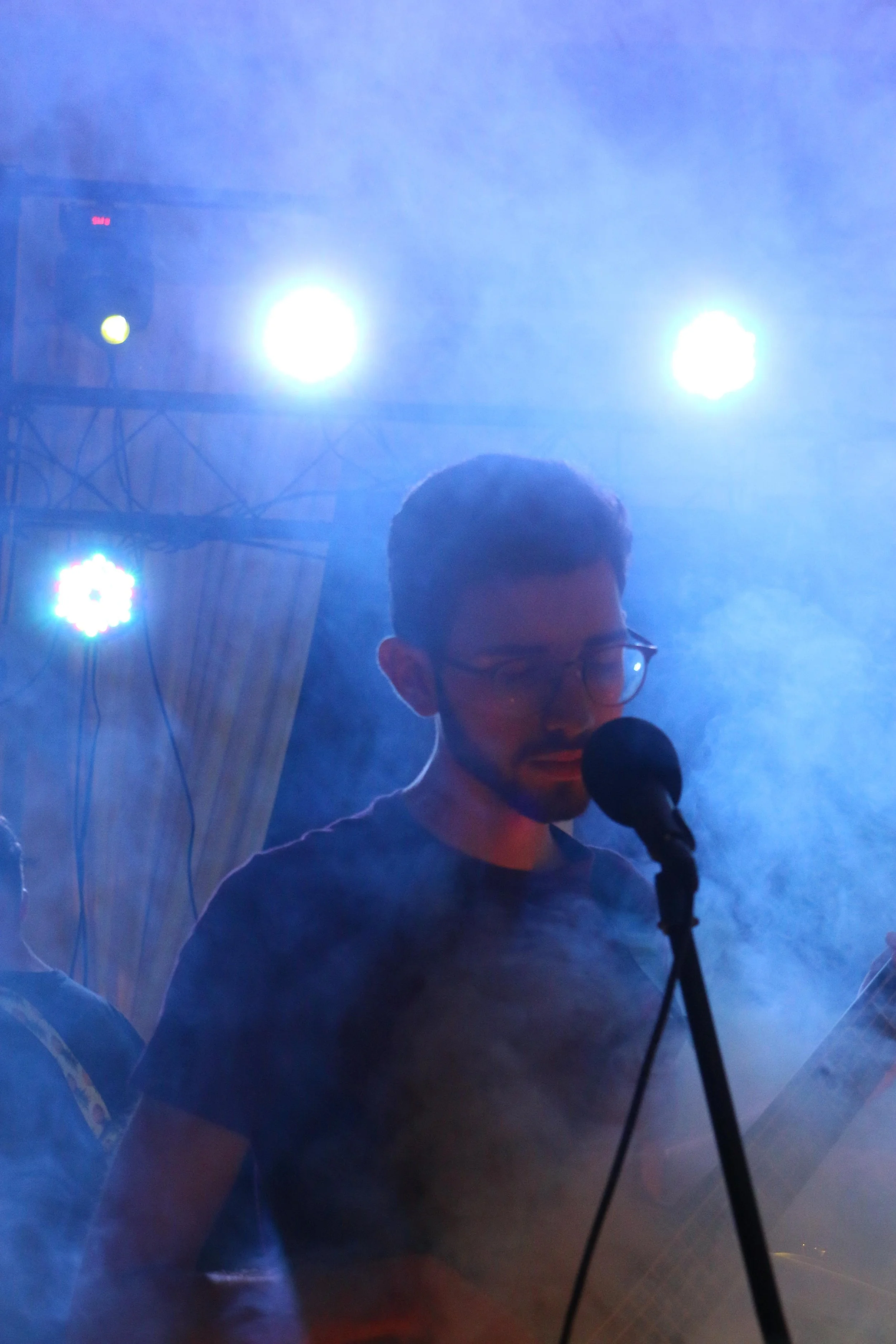 A man with glasses singing into a microphone on a dimly lit stage with blue stage lights and fog.