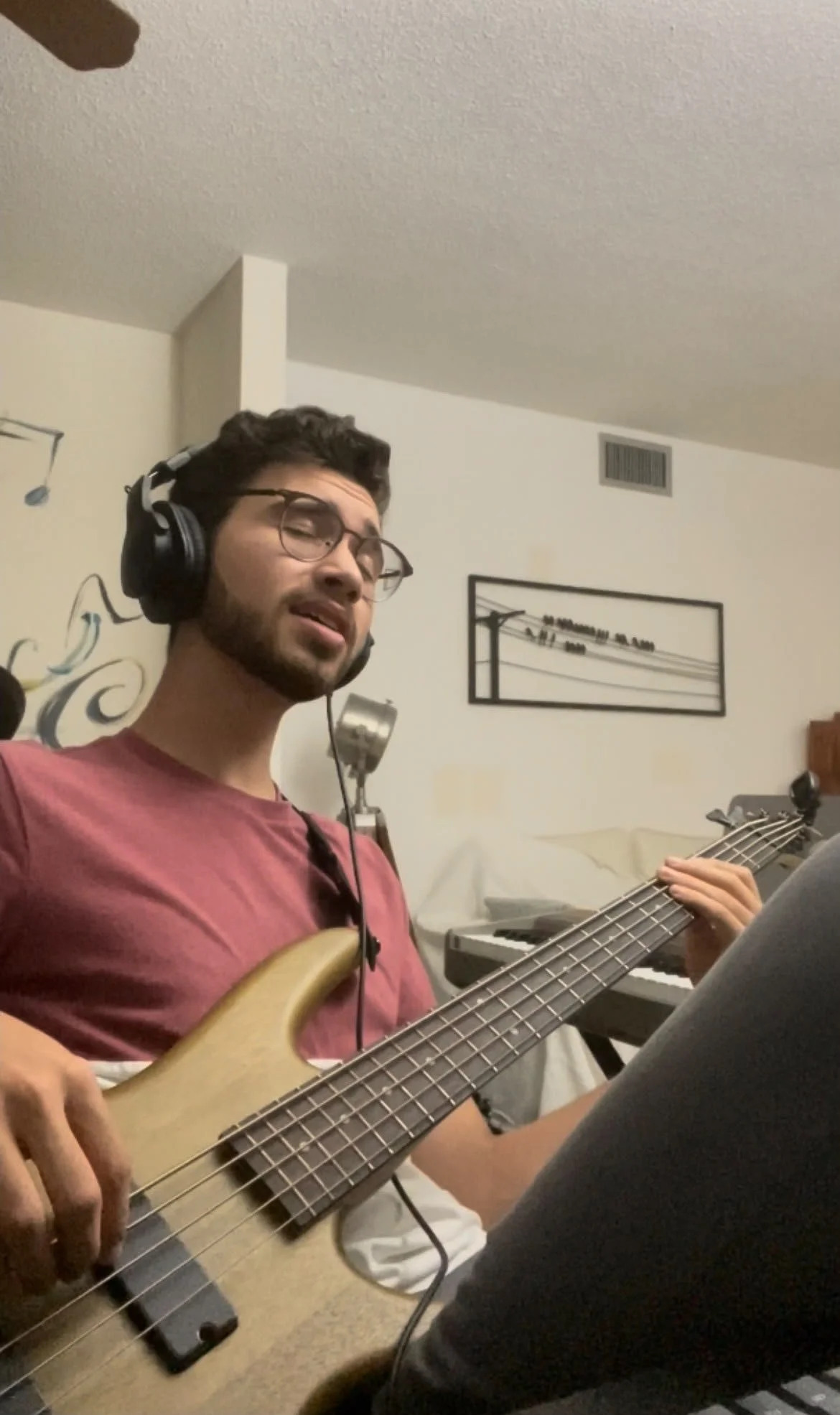 A young man wearing glasses and headphones, playing an electric bass guitar in a room with a keyboard, a wall decor, and a bed in the background.
