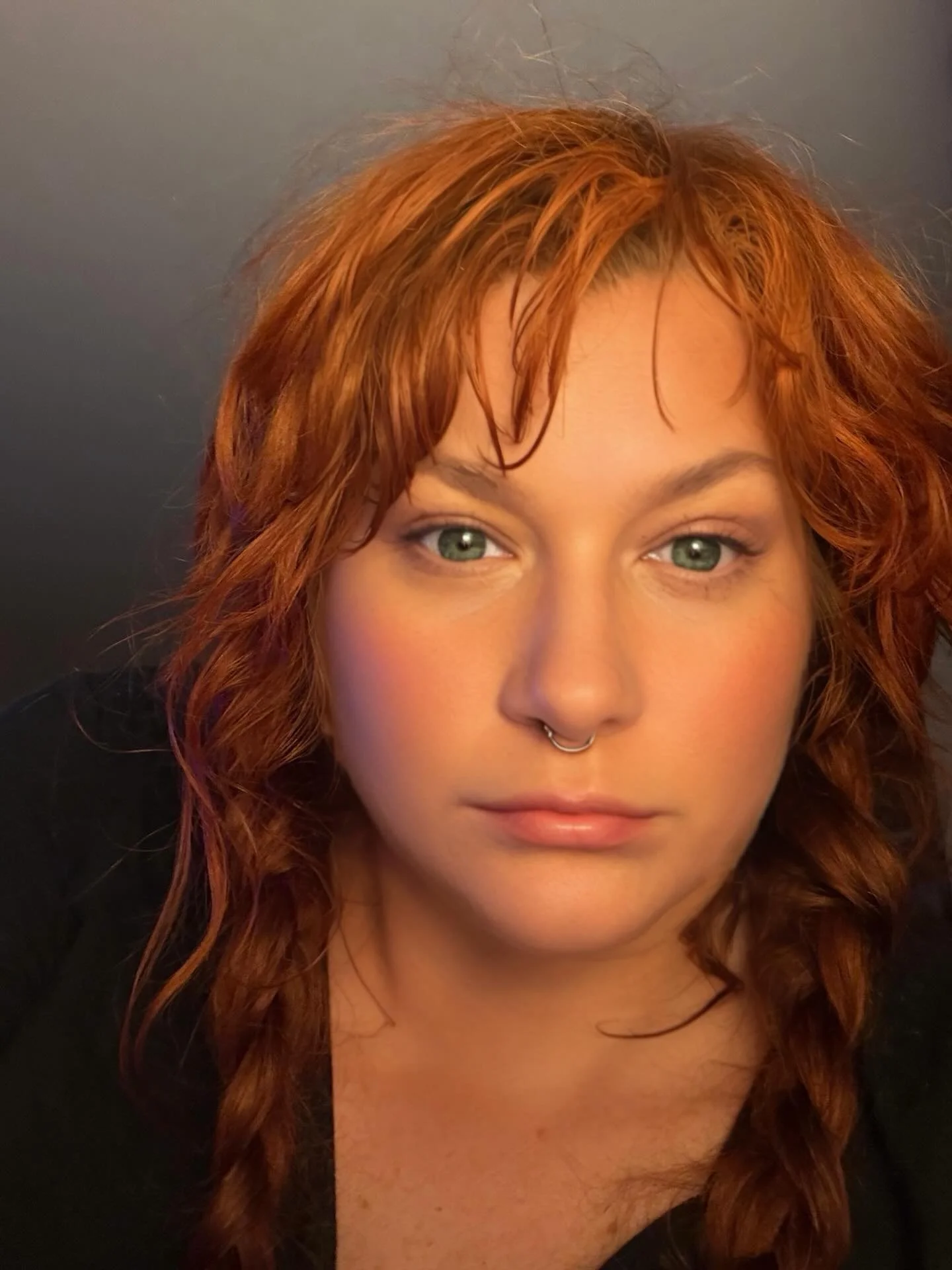 Close-up of a woman with curly auburn hair and green eyes, wearing a septum piercing, against a dark background.
