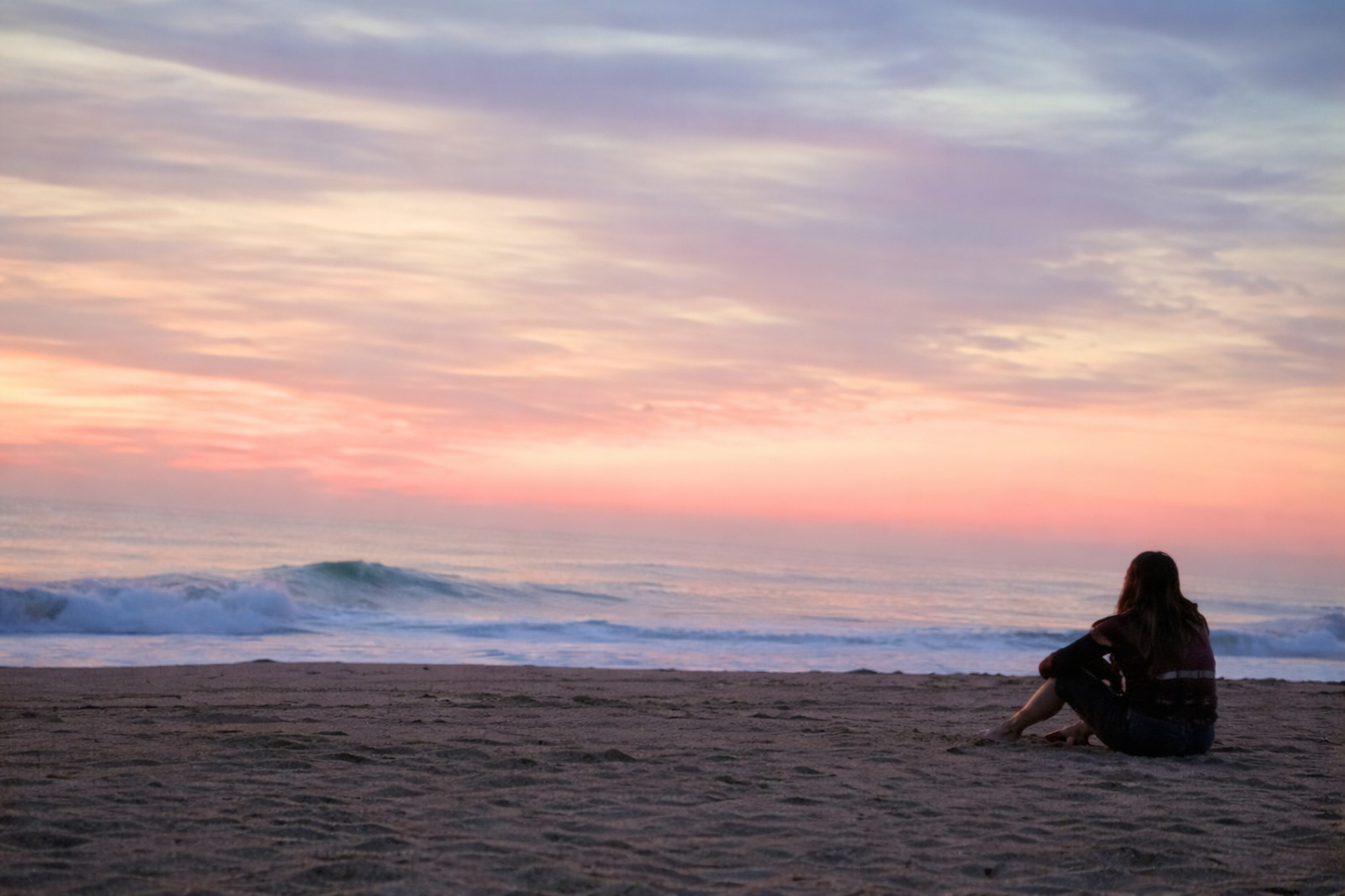 A person sitting on the sandy beach at sunset, gazing at the ocean with waves and pastel-colored sky.