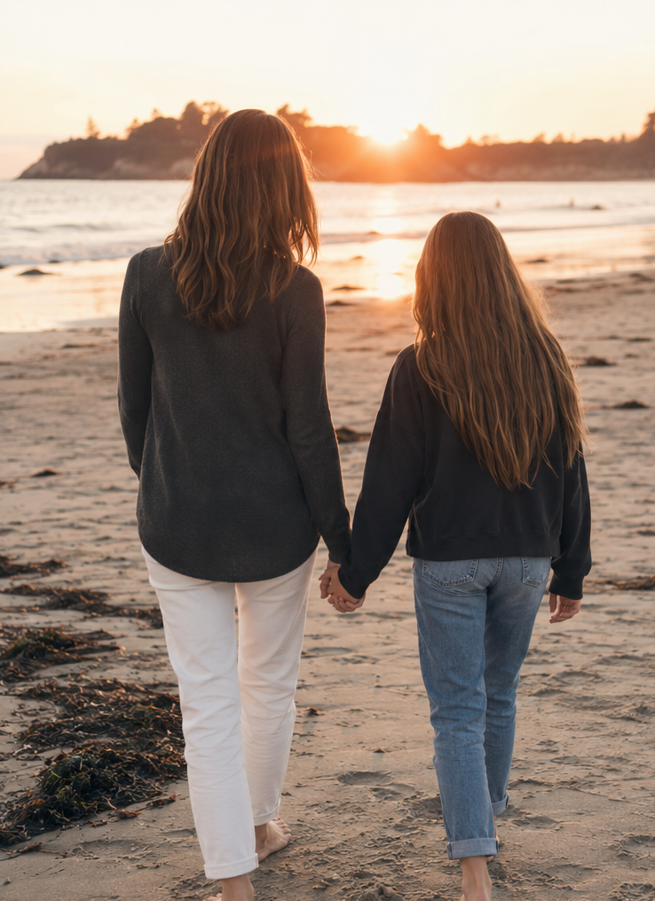 A woman and a girl holding hands walking on a beach at sunset.