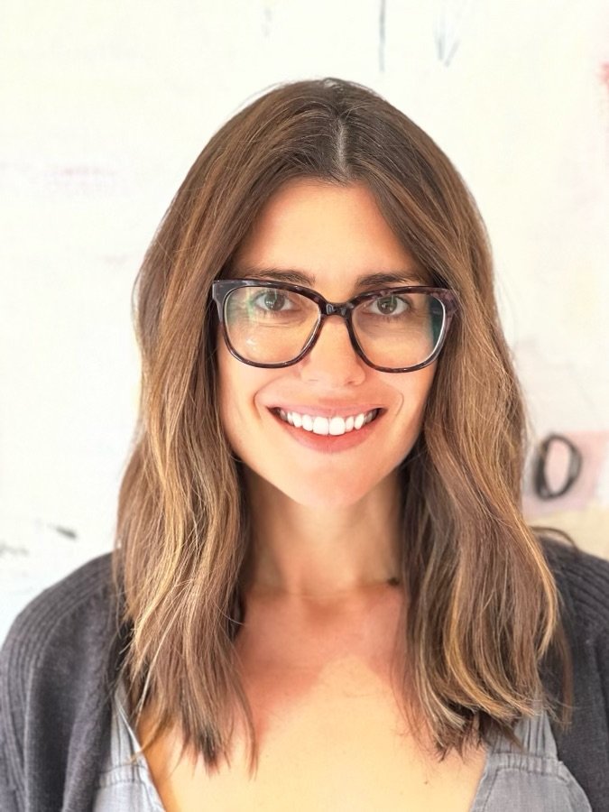 A woman with shoulder-length brown hair, glasses, and a gray top smiling at the camera.