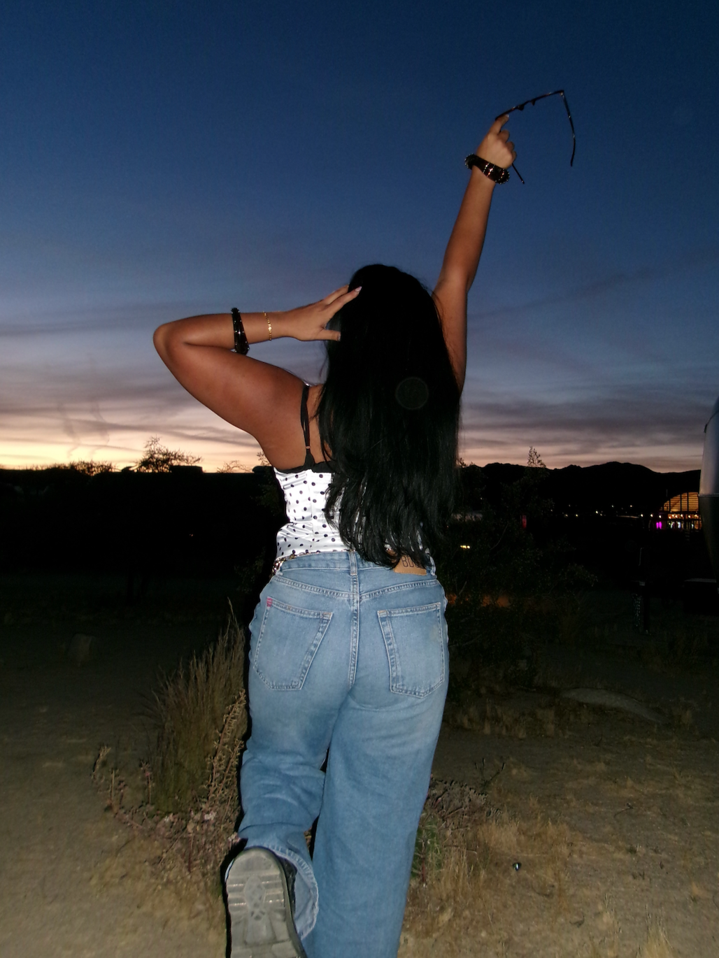 A woman standing outdoors at sunset, holding her sunglasses in one hand raised above her head, wearing a white polka dot top and jeans, with her other hand touching her head, against a backdrop of a colorful sky and distant hills.