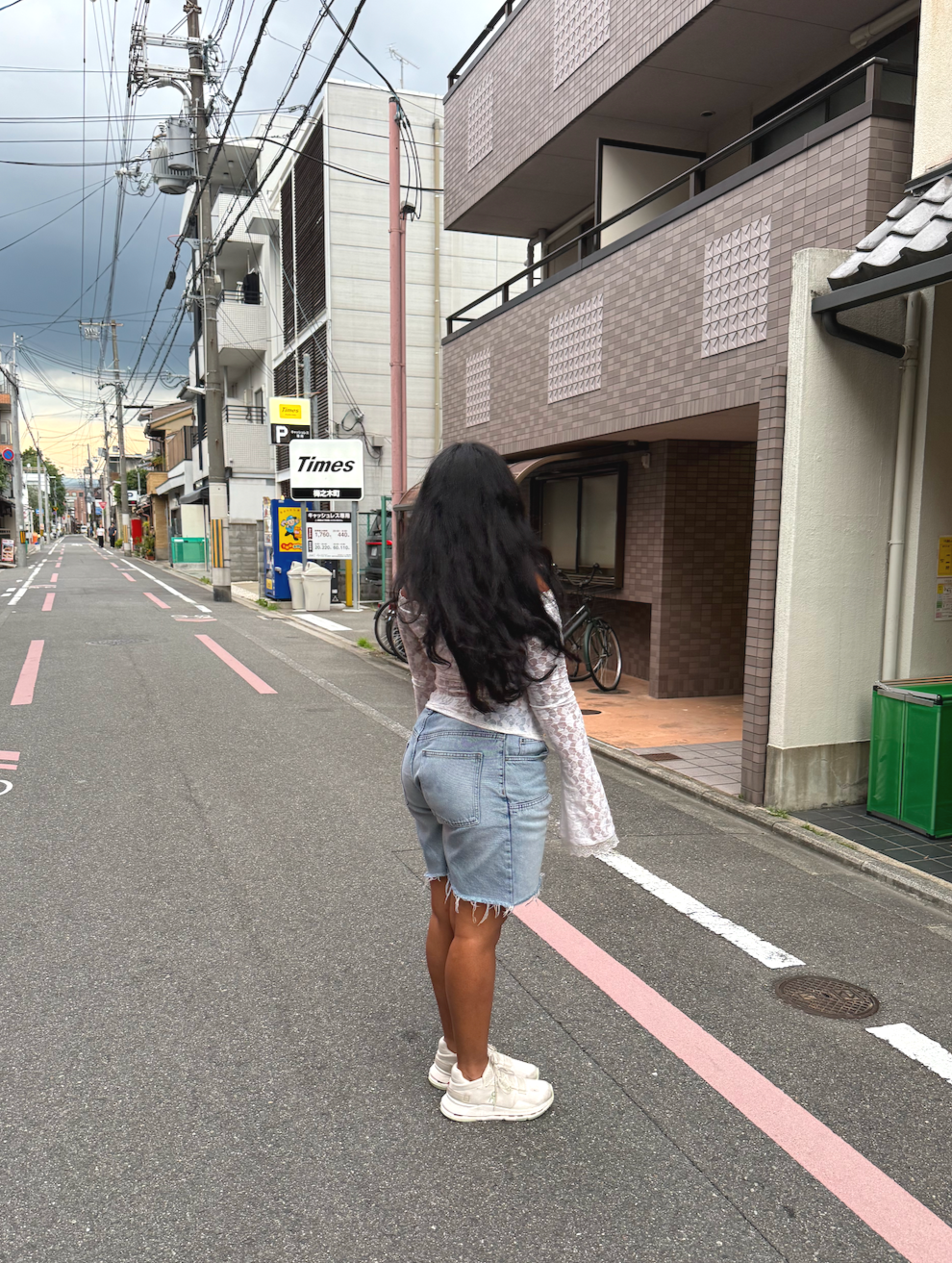 A woman with long black hair wearing a white lace long sleeve shirt, denim shorts, and white sneakers standing on a deserted city street during cloudy weather.