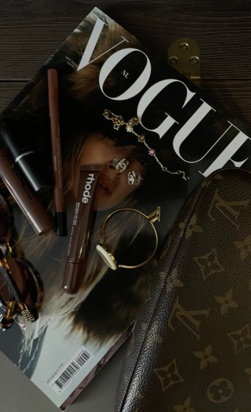 A Vogue magazine featuring a woman on the cover, several makeup products including lip liners or eyeliners, a gold bracelet, a pair of sunglasses, a Louis Vuitton handbag, and a gold jewelry chain with charms, all placed on a wooden surface.