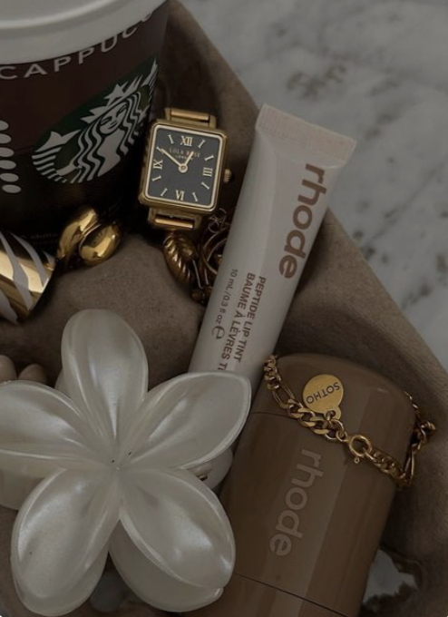 A collection of items including a Starbucks coffee cup, a black watch with gold accents, a white lip balm tube, a gold chain bracelet, a brown container with the brand name 'Rhode,' and a white ceramic flower-shaped dish.