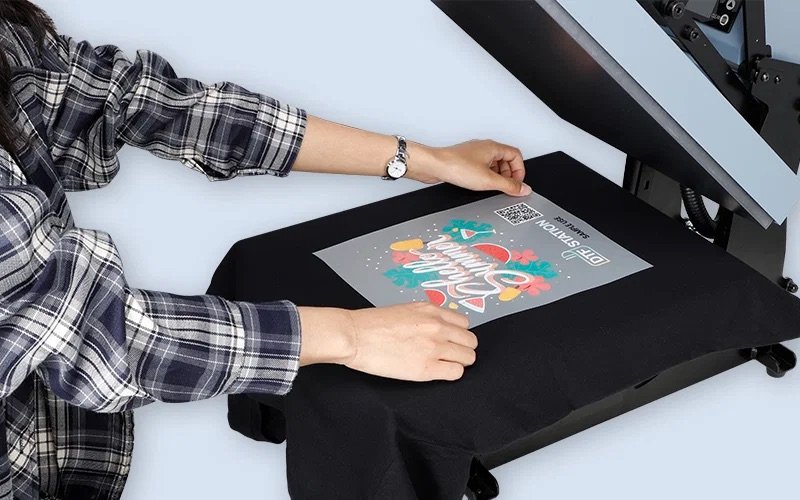 Person using a heat press machine to apply a colorful holiday-themed design onto a black fabric item.