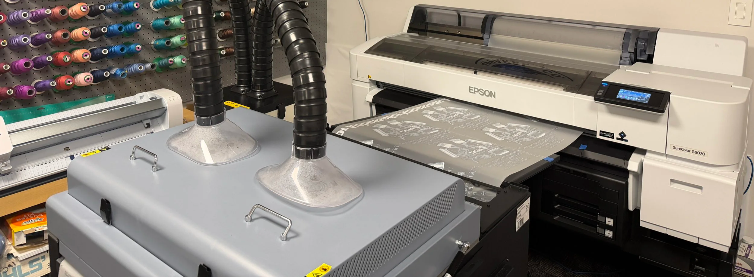 A large Epson printer printing a design sheet, with fabric and vent hoods in the foreground, and colored thread spools on a wall in the background.