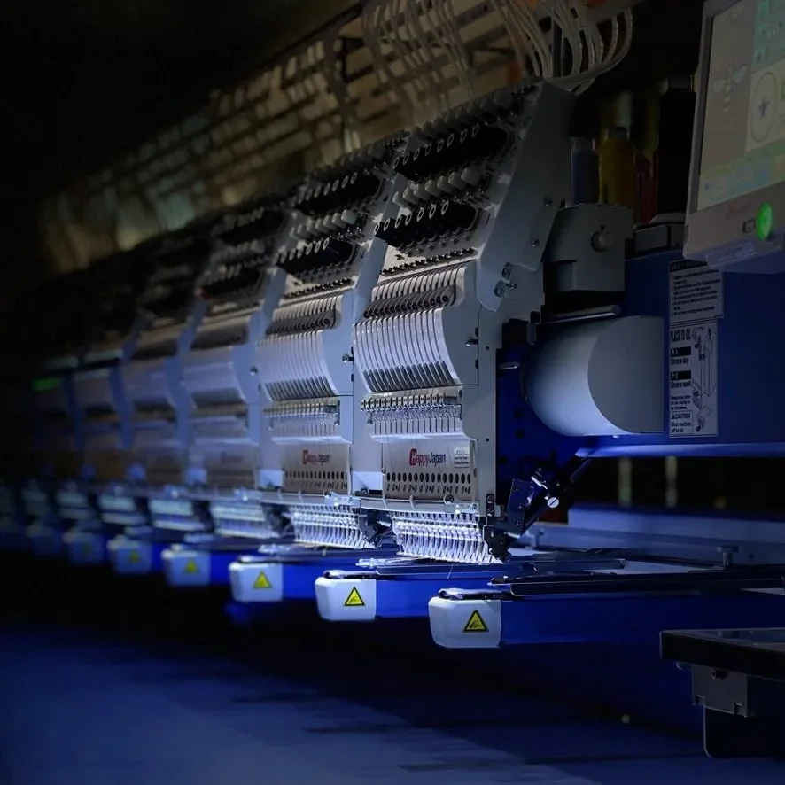 Close-up of industrial embroidery machines in operation, illuminated with blue light, with various components and wires visible.