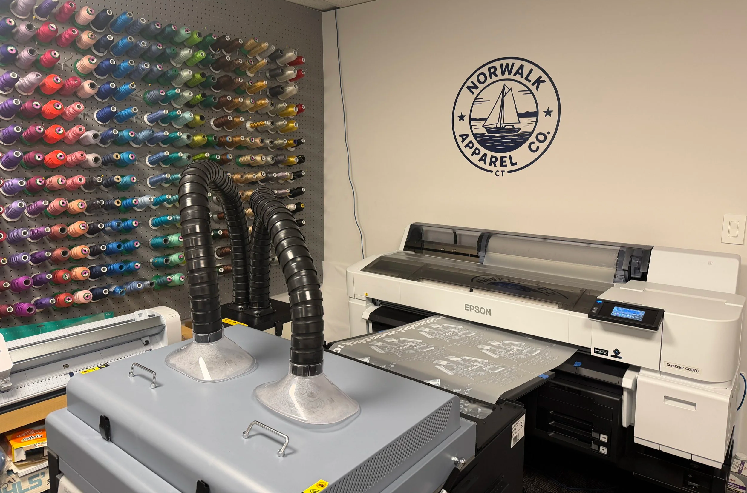 Wall with colorful spools of thread, printing equipment including a copier and printer, and an 'Norwalk Apparel Co.' logo with a sailboat on the wall.