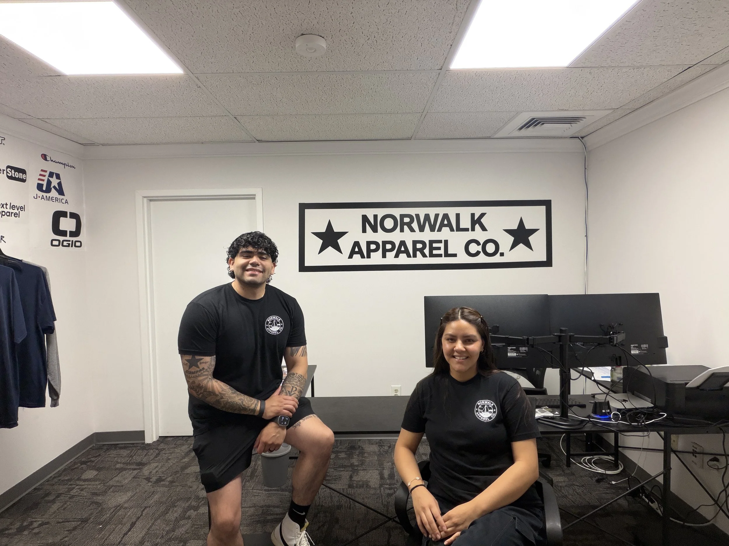 Two young people, a man and a woman, smiling and posing in an office. The man has curly hair, tattoos on his arms, and is wearing a black shirt and shorts. The woman has straight hair, is seated, and is wearing a black t-shirt with a white logo. There are office desks, computers, and a wall with a sign that says "Norwalk Apparel Co." in the background.