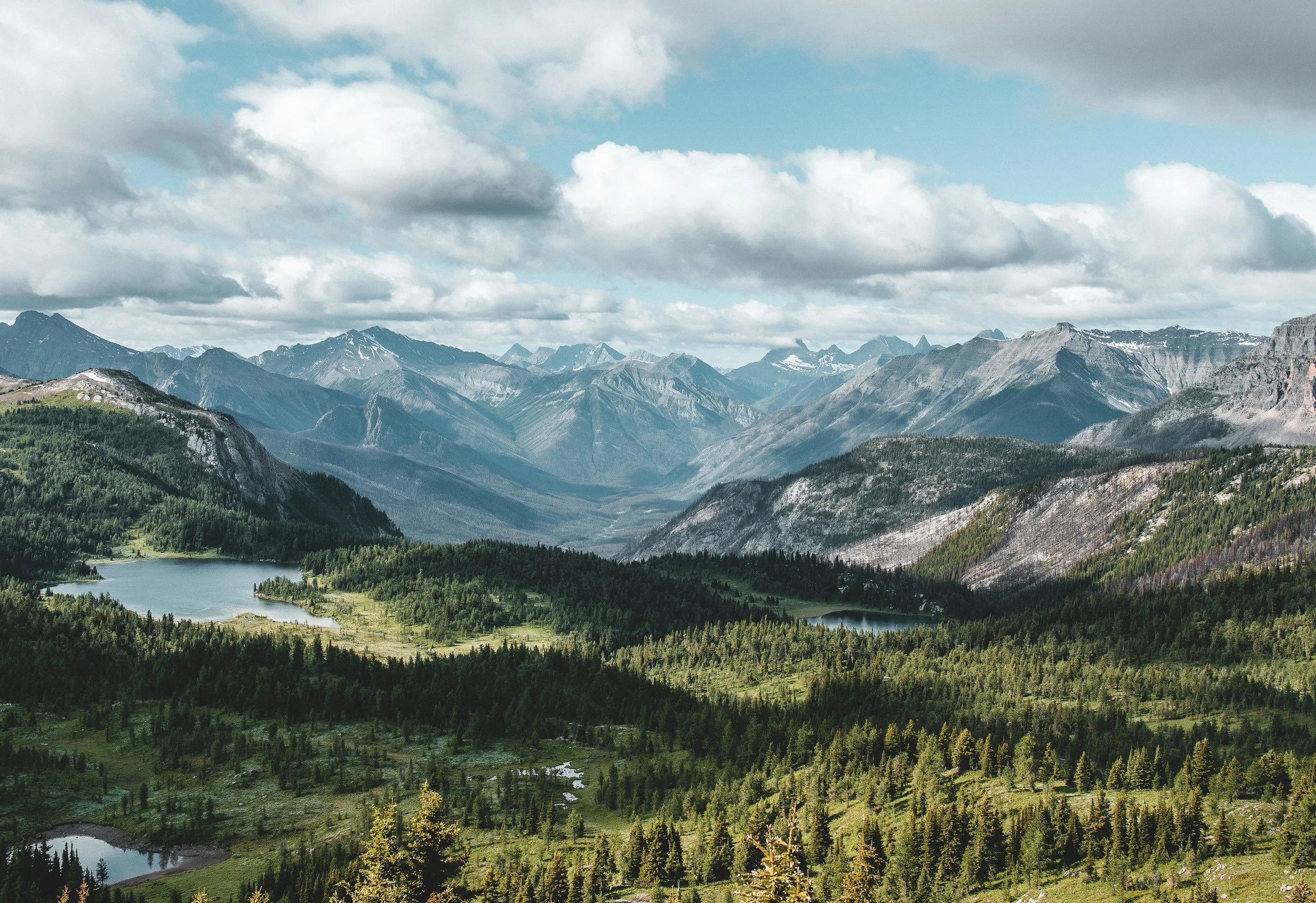 A panoramic view of a mountain range under a partly cloudy sky, with green forests and a small lake in the foreground.