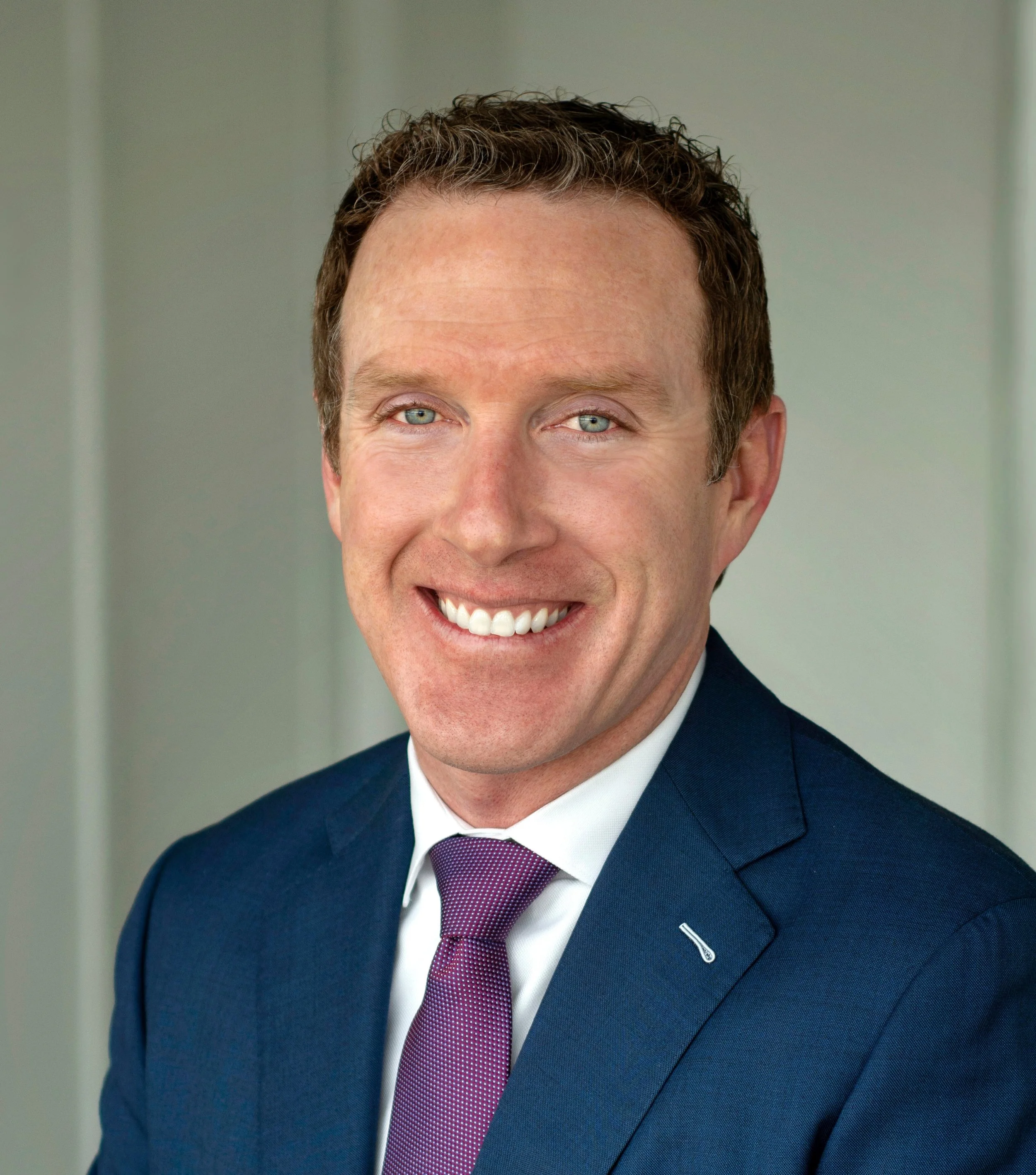 Close-up of a smiling man with blue eyes, wearing a navy suit, white shirt, and a purple tie with small white dots. The background is neutral and out of focus.