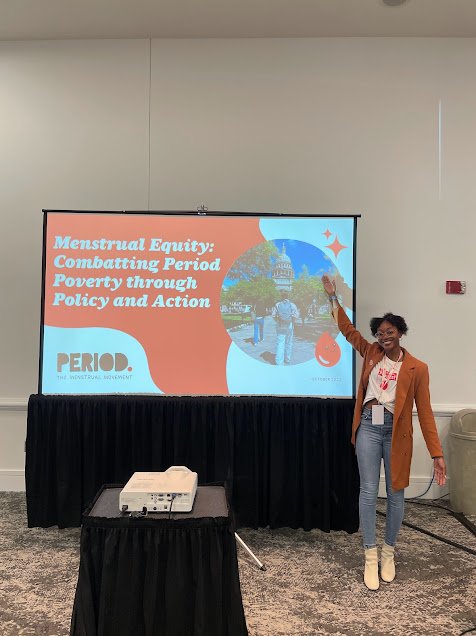 A woman standing next to a presentation slide on a large screen, pointing at the slide which features a cityscape, a logo, and text about menstrual equity and policy.