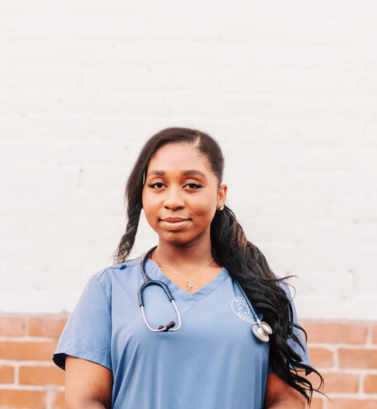 A young woman in professional medical scrubs with a stethoscope around her neck standing outdoors against a brick wall with a white background.