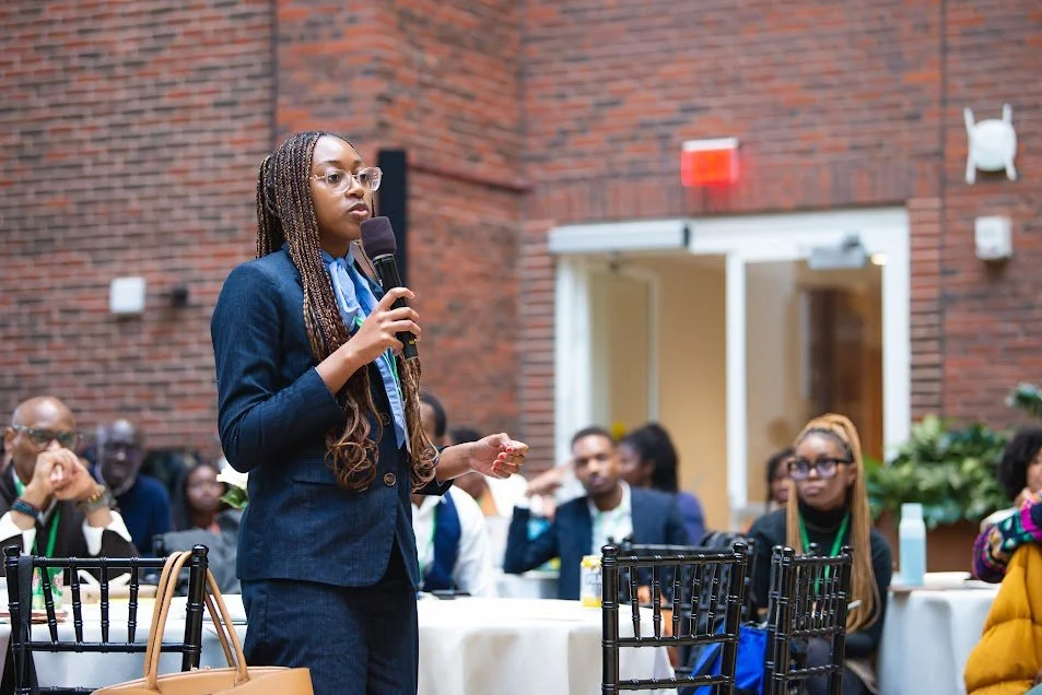 Woman in navy blazer speaking into microphone at conference, with seated audience listening.