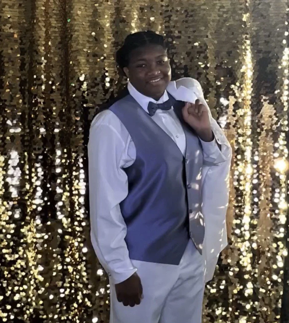 A person in a white dress shirt, gray vest, and bow tie standing in front of a gold sequin backdrop.