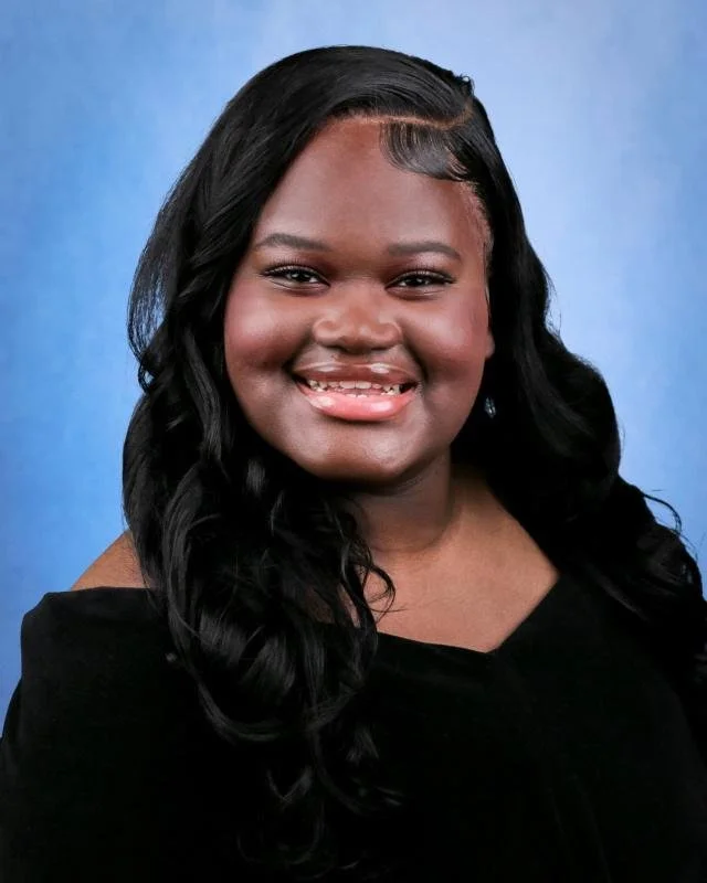 A young woman with long black curly hair, smiling, wearing a black dress, against a blue background.