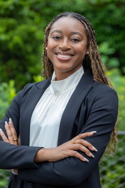 A smiling woman with long, braided hair wearing a dark blazer and white blouse, standing outdoors with green foliage in the background.
