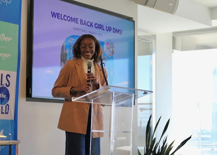 A woman standing at a clear podium, smiling, holding a microphone, with a large screen behind her displaying a welcoming message for a girls' summit.