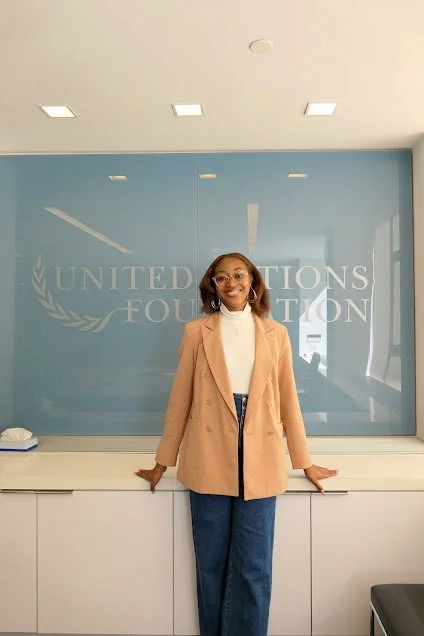 Woman standing in front of a glass wall with the United Nations Foundation logo, smiling and wearing a beige blazer over a white top with blue jeans.