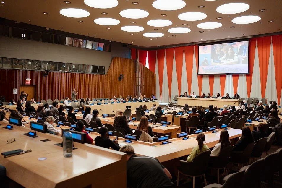 Group of people attending a conference or meeting in a large, modern auditorium with a large screen at the front and a panel of speakers.