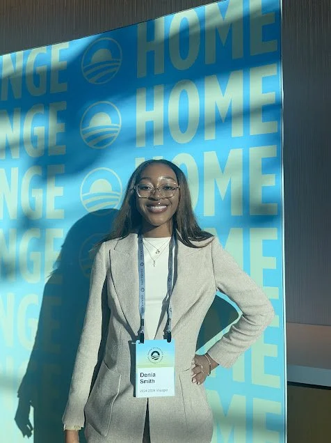 A woman wearing glasses and a beige blazer stands in front of a blue background with the words 'HOME' and a logo repeated, smiling at the camera.