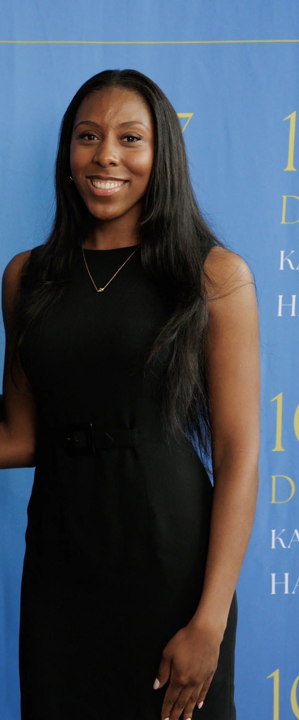 A young woman posing in front of a blue background with yellow lettering, wearing a black sleeveless dress and a gold necklace, smiling at the camera.