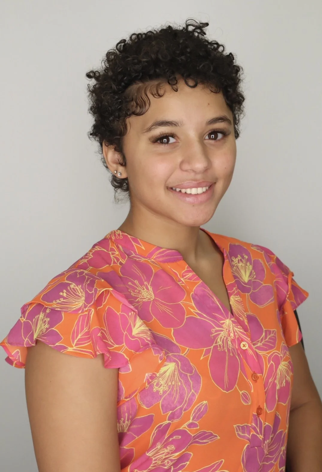 A young woman with short curly dark hair, wearing a colorful floral blouse in pink and orange, smiling at the camera against a plain light background.