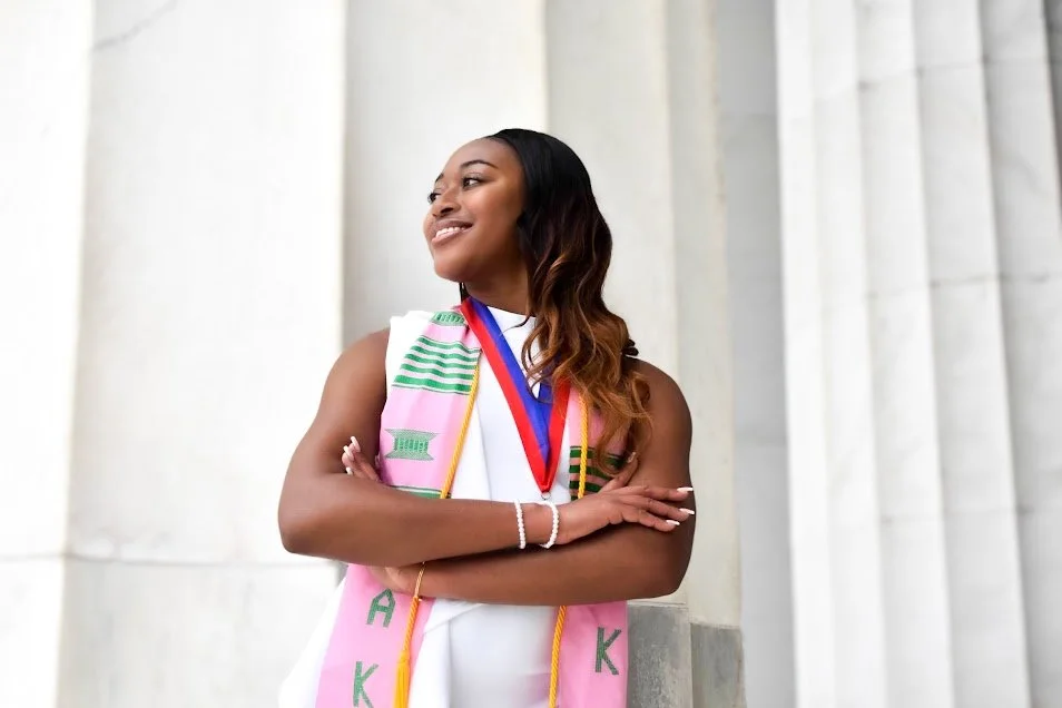 A young woman in a white dress standing outdoors in front of white columns, wearing a graduation stole and medal, smiling confidently with her arms crossed.