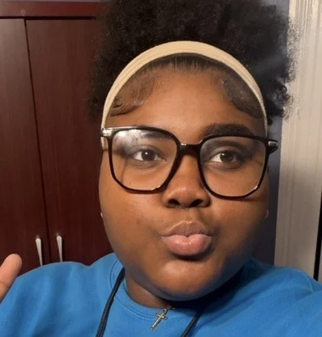 A young woman with glasses and a beige headband making a kissy face for the camera.