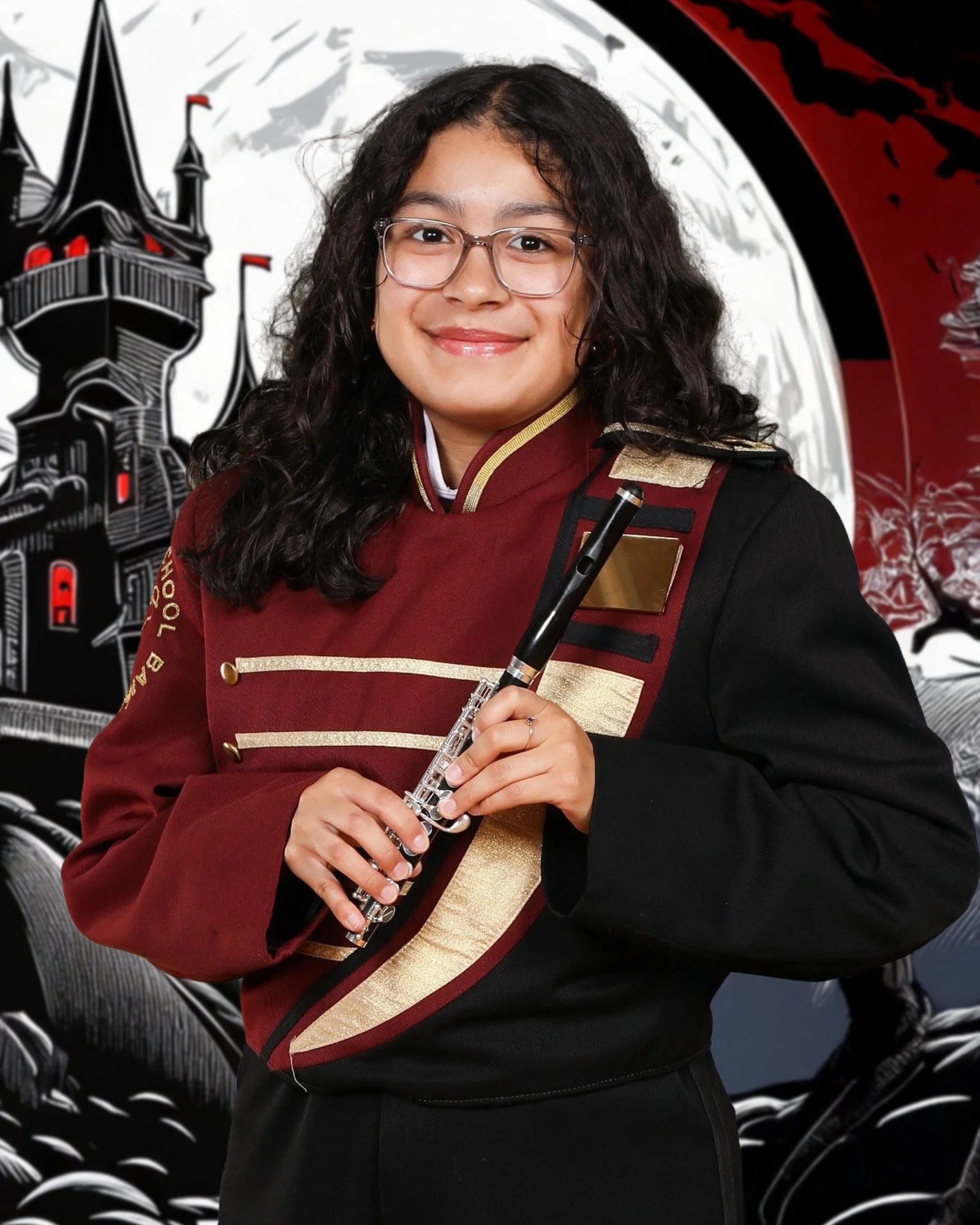A woman with dark curly hair, glasses, and a bright smile, holding a flute, dressed in a black and red academic or formal uniform with gold details, standing in front of a large black and white mural depicting a castle and buildings.