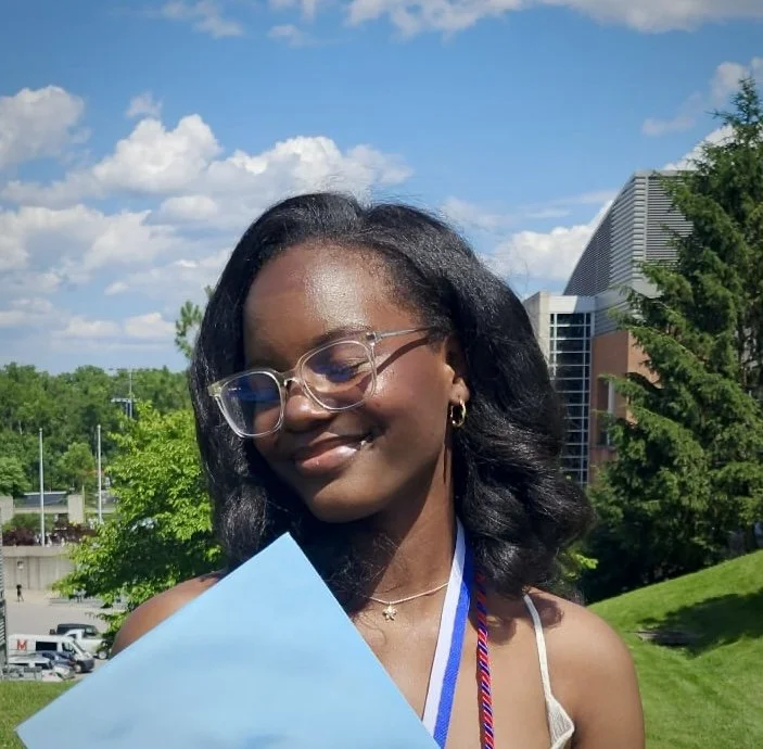 A young woman with dark hair and glasses holding a blue diploma outdoors on a sunny day, with trees and a modern building in the background.