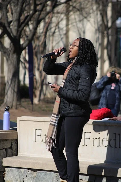 A woman with glasses and curly hair singing into a microphone outdoors during daytime, holding a phone in her other hand, with a person taking a photo in the background.