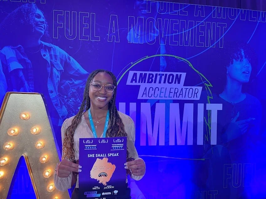 A woman with glasses and long braids smiling at an event, holding a purple certificate that says 'She Shall Speak.' Behind her is a blue-lit background with the text 'Full a Movement,' 'Ambition Accelerator,' and 'Summit,' along with a large decorative letter A with lights.
