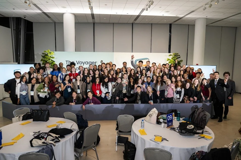 A large group of people smiling and posing for a photo in a conference room, with round tables set up in front and a presentation screen behind them that reads 'The Voyager.'