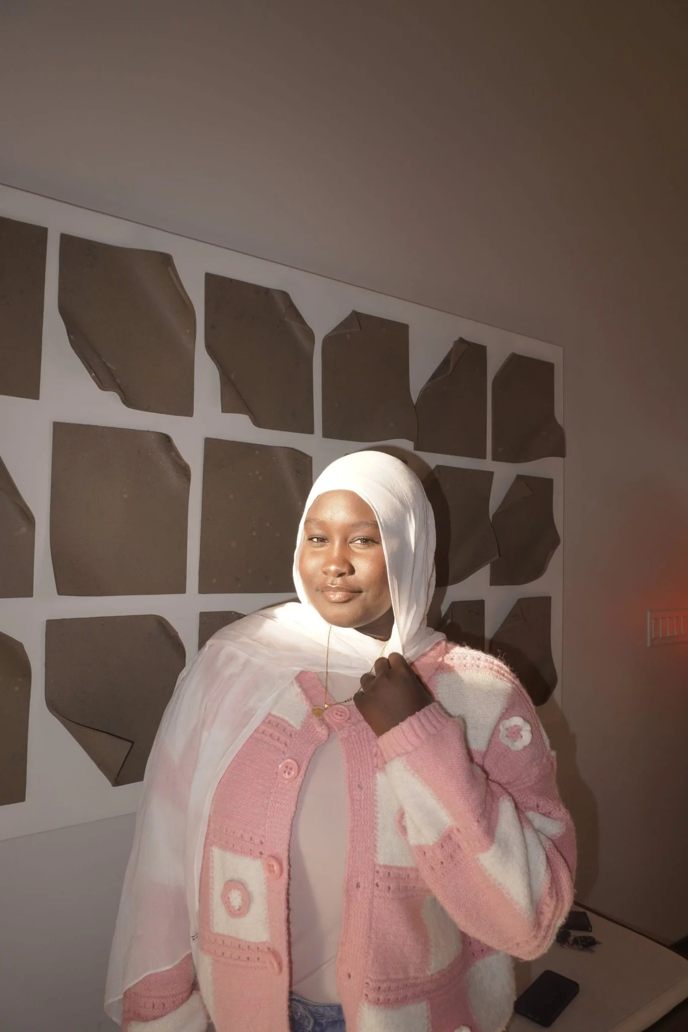 A woman wearing a white hijab and pink and white sweater standing in front of a wall with irregularly shaped dark panels.