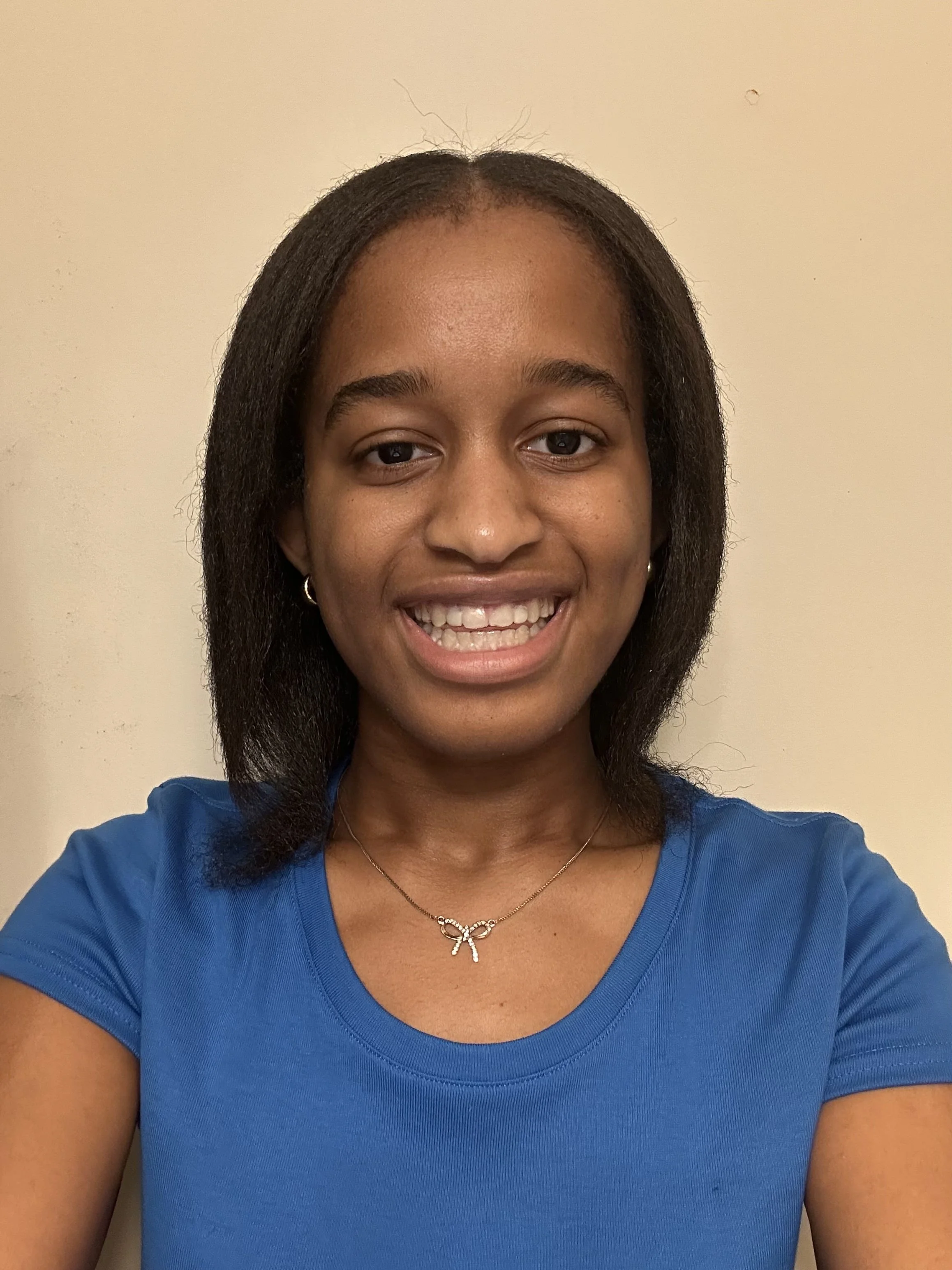 Close-up of a smiling woman wearing a blue top, a butterfly necklace, and small hoop earrings, with a plain beige wall in the background.