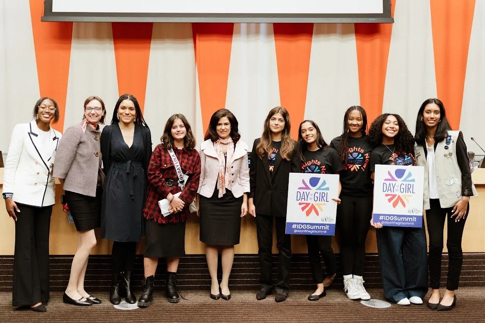 Group of nine women standing together in front of a stage, some holding signs for 'DAY of GIRL' and '#IDGSummit' at an event.